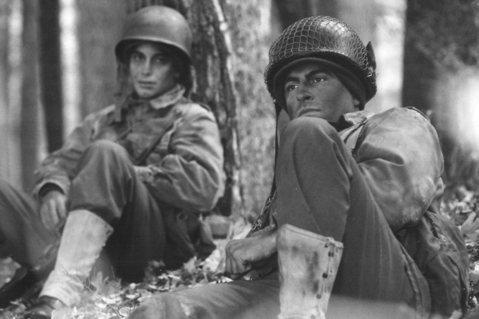 Two American soldiers resting against a tree in the woods, waiting